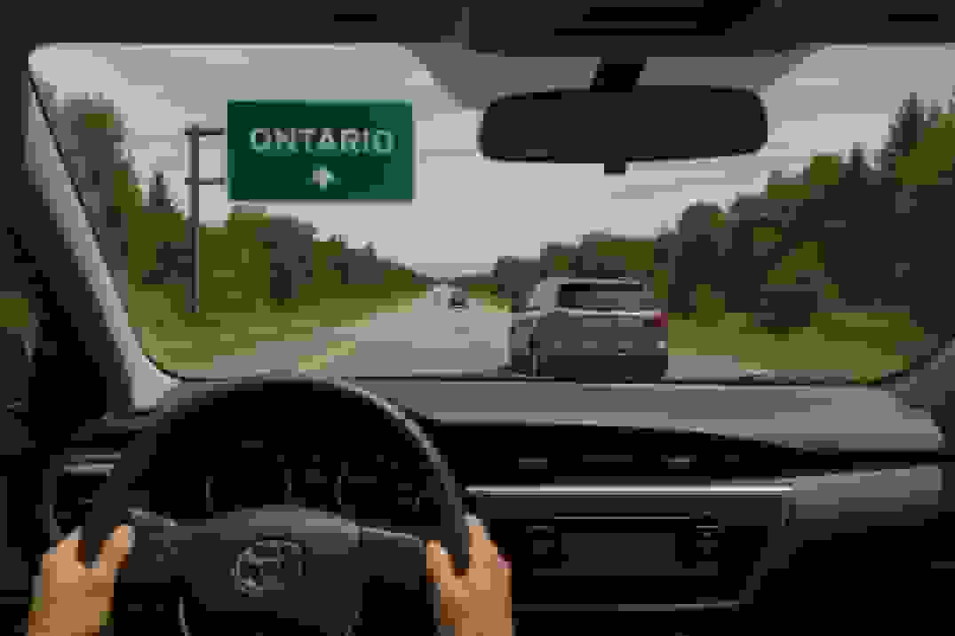 Driver’s view from inside a car on a two-lane highway approaching an overhead green sign reading ‘ONTARIO’, with a silver SUV ahead and tree-lined shoulders under a partly cloudy sky.
