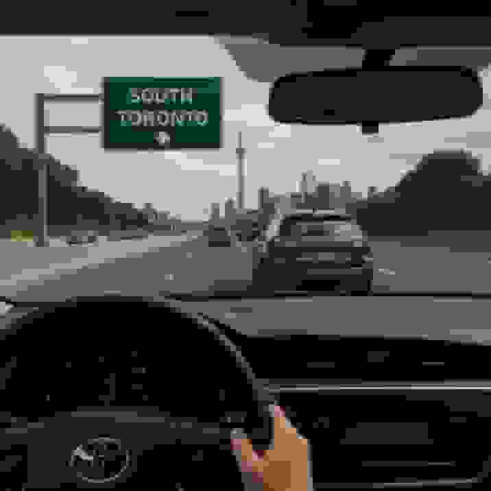 Driver’s perspective from behind the steering wheel on a multi-lane highway under a green ‘SOUTH TORONTO’ sign, with a silver crossover ahead and the CN Tower and downtown skyline visible in the distance.