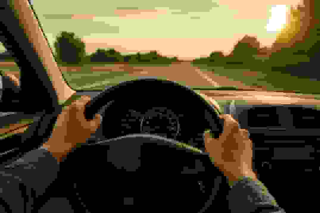 View from inside a car at sunset with hands on the steering wheel, looking out at an empty road flanked by trees bathed in warm golden light.