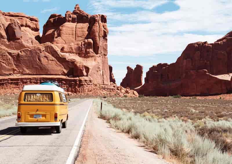 Yellow Volkswagen camper van driving through a red rock canyon desert road under a clear blue sky.