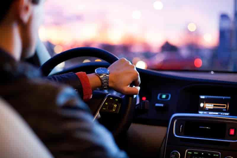Driver's hands gripping a steering wheel at night with blurred city lights glowing in the background.